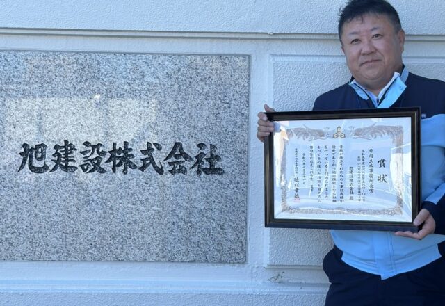 日向土木事務所長賞の賞状を手にする旭建設の社員。本社の社名板の前にて