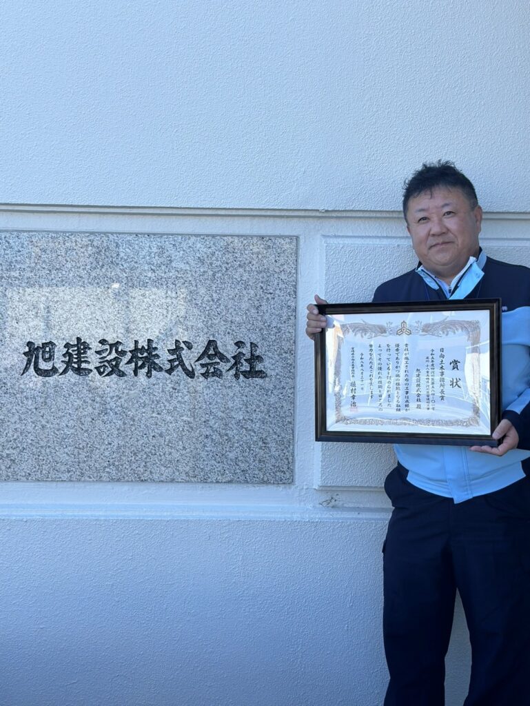 日向土木事務所長賞の賞状を手にする旭建設の社員。本社の社名板の前にて