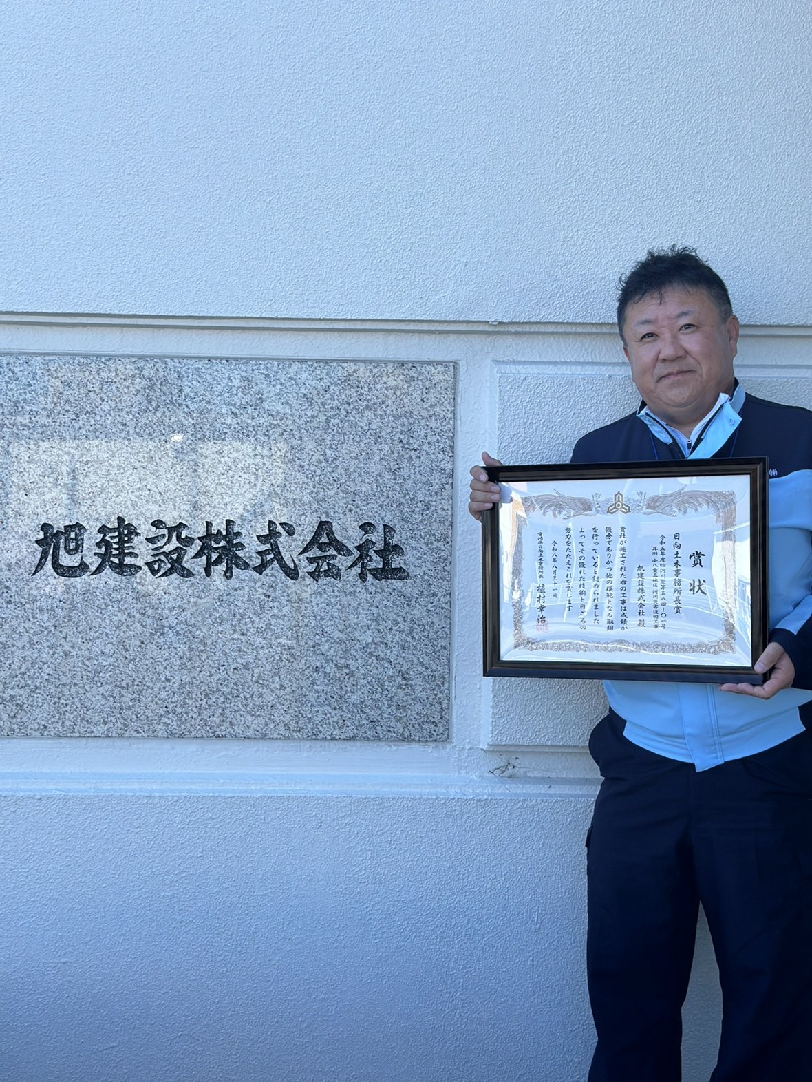 日向土木事務所長賞の賞状を手にする旭建設の社員。本社の社名板の前にて