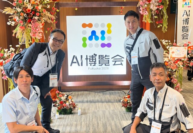 AI博覧会 Fukuoka 2026 の会場入口のロゴの前で、参加した社員4名